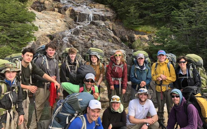 A group of people wearing backpacks pose for a photo in front of a small waterfall.